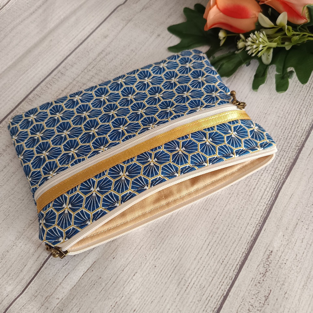 Neceser plano de tela azul con dos cremalleras y detalles dorados en una mesa de madera blanca junto a flores secas"