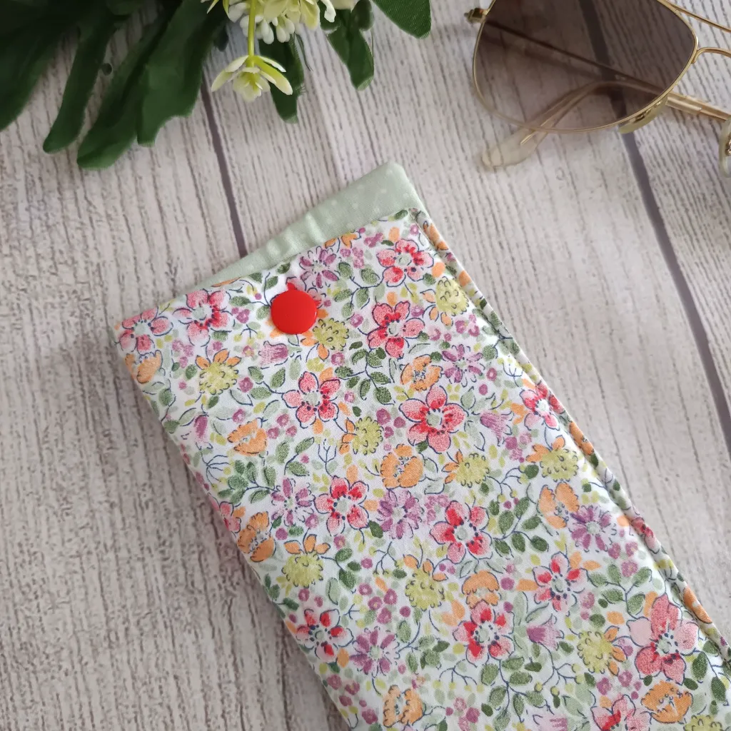 Funda de gafas artesanal hecha a mano en tela de algodón con estampado floral multicolor en rosa, naranja, lila y verde sobre fondo blanco, con cierre a presión rojo, fotografiada sobre madera clara junto a unas gafas de sol doradas